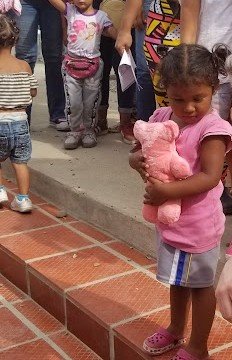 A young girl hugging a pink teddy bear, standing in a crowd of women and children at a Hope 143 distribution.